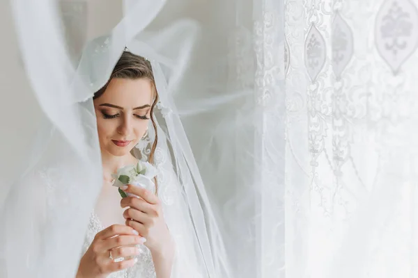 The bride is dressed in an elegant dress, covered with a veil, posing and holding a boutonniere. Wedding photo, morning of the bride
