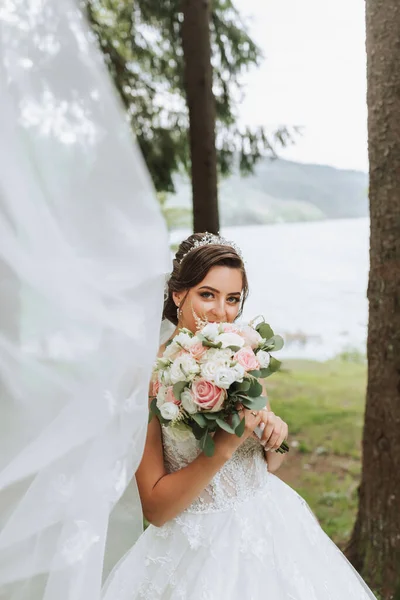 Portrait of a bride with a bouquet against the background of tall trees and a lake. A beautiful young bride is holding a wedding bouquet in her hands, the bride\'s veil is blowing in the wind.