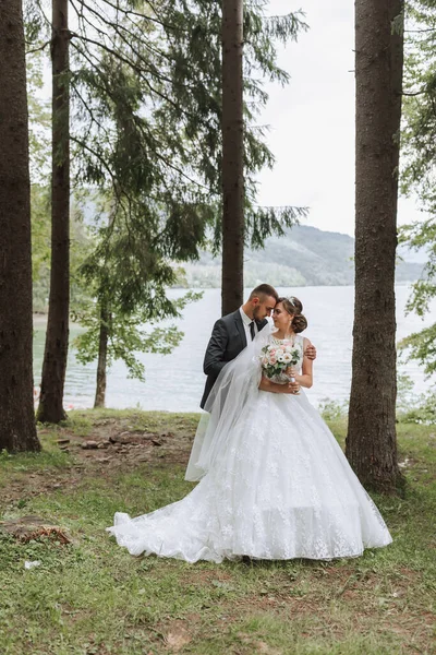A wedding couple is enjoying the best day of their lives against the backdrop of a lake and tall trees. The groom hugs the bride.
