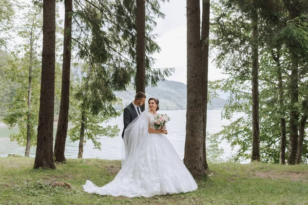 A wedding couple is enjoying the best day of their lives against the backdrop of a lake and tall trees. The groom hugs the bride from behind