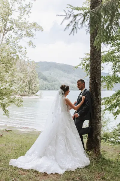 A wedding couple is enjoying the best day of their lives against the backdrop of a lake and tall trees. The groom hugs the bride.