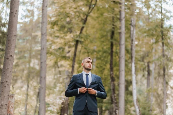 森や公園の背の高い木の背景に対する古典的なスーツの若い男 グロムは彼のジャケットを締める — ストック写真