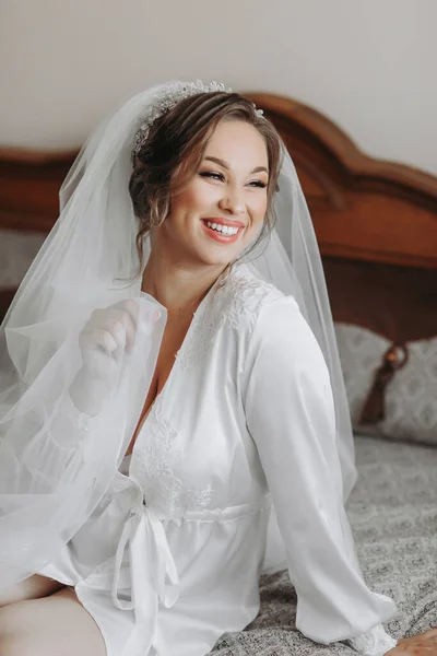 Beautiful young bride in white petticoat and tiara sitting posing on bed in her bedroom. Final preparations for the wedding. The bride is waiting for the groom. Morning, bride.