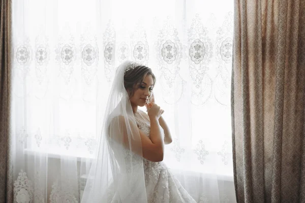 Portrait of the bride. The bride is dressed in an elegant dress, posing by the window wearing earrings. Tiara and jewelry. Open bust. Morning of the bride