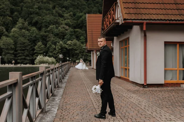 結婚式の肖像画 黒いスーツを着た男がブーケで橋の上に立って見つめている ファイアンス マスカリン ビジネススタイル — ストック写真