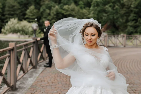 Wedding portrait. The bride in a white dress on the bridge with a flowing veil, the groom is standing behind her. Sincere smile. Wind and veil. Diadem.