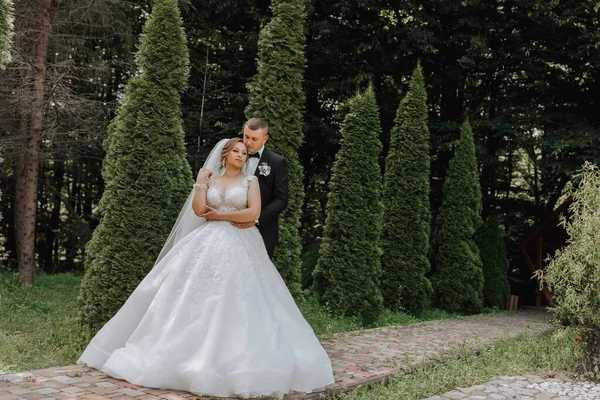 Wedding portrait. The bride in an elegant dress stands in front of the groom in a classic suit, against the background of green trees. Gentle touch. Summer wedding. A walk in nature