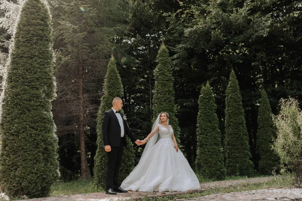 Wedding portrait.The bride and groom are holding hands. The bride in an elegant dress, the groom in a classic suit against the background of green trees. Gentle touch. Summer wedding. A walk in nature