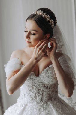 Preparation for the wedding. Beautiful young bride in white luxurious wedding dress, tiara on head, long veil in royal hotel room. A luxurious model clings to an earring by the window clipart