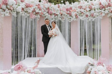 Düğün günlerinde gelin ve damat, pembe ve beyaz çiçeklerden yapılmış bir kemerle seremonide. Güzel yeni evliler, uzun trenli beyaz elbiseli genç bir kadın, siyah takım elbiseli adamlar..