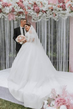 Düğün günlerinde gelin ve damat, pembe ve beyaz çiçeklerden yapılmış bir kemerle seremonide. Güzel yeni evliler, uzun trenli beyaz elbiseli genç bir kadın, siyah takım elbiseli adamlar..