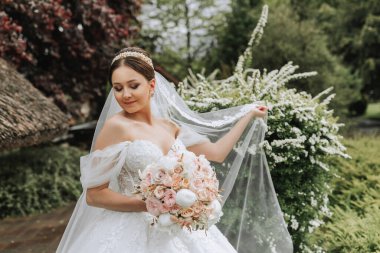 Beyaz gelinlik içinde güzel bir gelinin portresi. Uzun bir tren, modern saç stili ve bahçede yürüyen bir duvak. Düğün konsepti