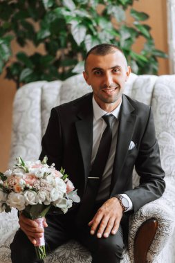 Elinde bir buket çiçekle şık bir damat pahalı bir otel odasında bir sandalyede oturuyor. Damat sabahı. Damat düğün töreninden önce hazırlanıyor.