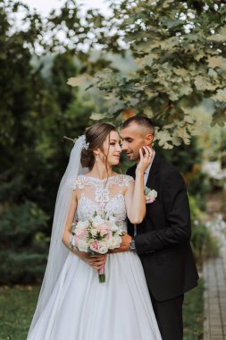 Modaya uygun damat ve beyaz elbiseli yakışıklı esmer gelin profesyonel makyaj ve çiçek buketi ile sarılıyor, parkta, bahçede, ormanda gülüyor. Düğün fotoğrafçılığı