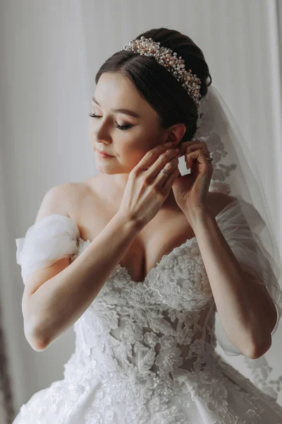 Preparation for the wedding. Beautiful young bride in white luxurious wedding dress, tiara on head, long veil in royal hotel room. A luxurious model clings to an earring by the window