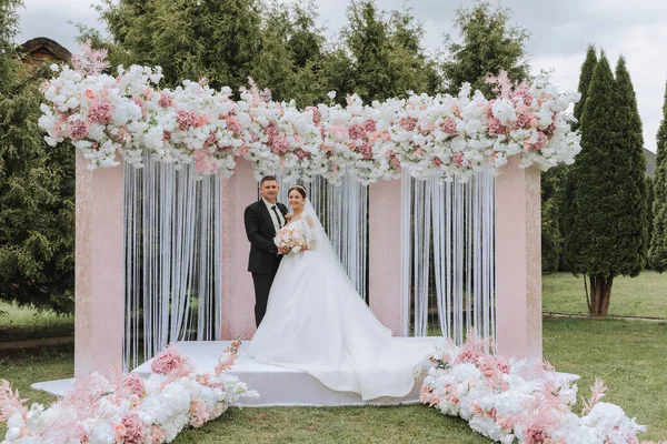 ピンクと白の花で作られたアーチで 結婚式の日に魅力的な花嫁とグルーミング 美しい新婚夫婦 長い列車の白いドレスの若い女性 黒いスーツの男性 — ストック写真