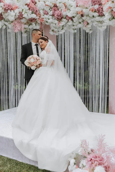 Attractive bride and groom at the ceremony on their wedding day with an arch made of pink and white flowers. Beautiful newlyweds, a young woman in a white dress with a long train, men in a black suit.