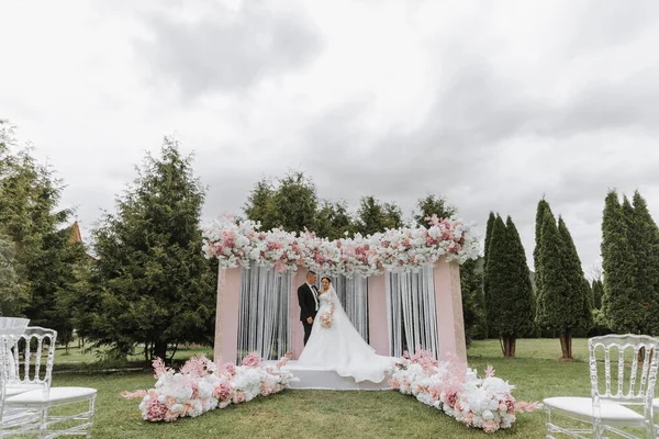 Attractive bride and groom at the ceremony on their wedding day with an arch made of pink and white flowers. Beautiful newlyweds, a young woman in a white dress with a long train, men in a black suit.