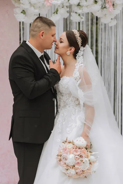 Attractive bride and groom at the ceremony on their wedding day with an arch made of pink and white flowers. Beautiful newlyweds, a young woman in a white dress with a long train, men in a black suit.