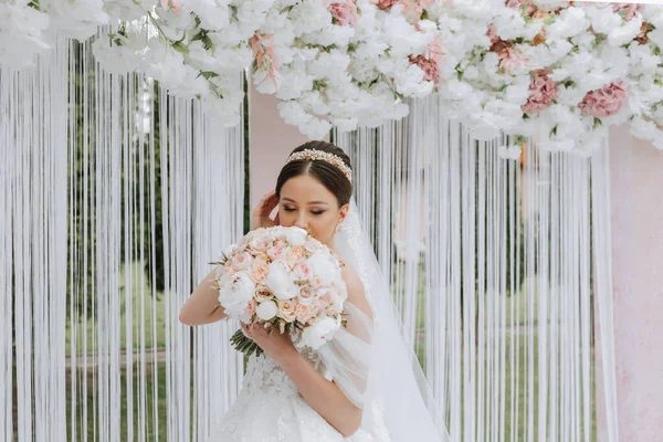 Attractive bride at the ceremony on her wedding day with an arch made of pink and white flowers. Beautiful newlyweds, a young woman in a white dress with a long train, men in a black suit.