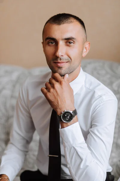 A stylish groom with a watch on his hand and a tie is sitting on a chair in an expensive hotel room. Groom\'s morning. The groom is getting ready in the morning before the wedding ceremony.