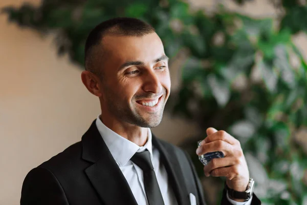A stylish groom with a bouquet of flowers sprays himself with perfume. Groom\'s morning. The groom is getting ready in the morning before the wedding ceremony