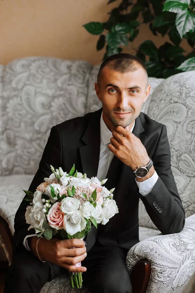 A stylish groom with a bouquet of flowers is sitting on a chair in an expensive hotel room. Groom\'s morning. The groom is getting ready in the morning before the wedding ceremony