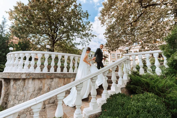 一个穿着白衣的年轻新娘和一个穿着西服的黑发新郎走在一座古城公园的台阶上的画像 美丽而浪漫的婚礼 幸福的一对 — 图库照片