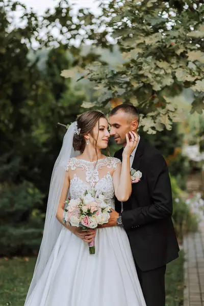 Fashionable groom and cute brunette bride in white dress with professional make-up and bouquet of flowers are hugging, laughing in park, garden, forest outdoors. Wedding photography