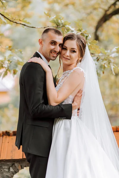 Fashionable groom and cute brunette bride in white dress with professional make-up and bouquet of flowers are hugging, laughing in park, garden, forest outdoors. Wedding photography