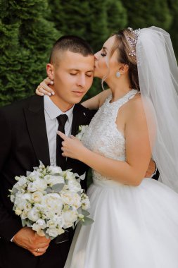 Siyah takım elbiseli şık bir damat ve uzun peçeli beyaz elbiseli şirin bir gelin eski bir evin yakınındaki bir parkta sarılıyorlar. Gülümseyen ve mutlu yeni evlilerin düğün portresi.