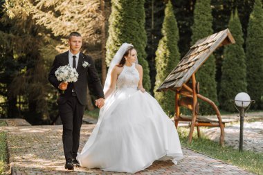 Siyah takım elbiseli şık bir damat ve uzun peçeli beyaz elbiseli şirin bir gelin yeşil uzun ağaçların yanında sarılıp yürüyorlar. Gülümseyen ve mutlu yeni evlilerin düğün portresi.