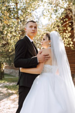 Siyah takım elbiseli şık bir damat ve uzun peçeli beyaz elbiseli şirin bir gelin eski bir evin yakınındaki bir parkta sarılıyorlar. Gülümseyen ve mutlu yeni evlilerin düğün portresi.