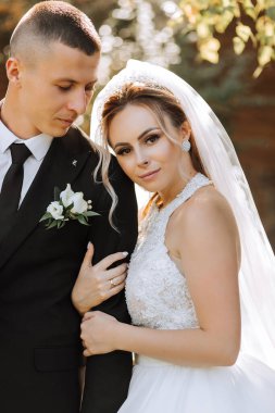 Siyah takım elbiseli şık bir damat ve uzun peçeli beyaz elbiseli şirin bir gelin eski bir evin yakınındaki bir parkta sarılıyorlar. Gülümseyen ve mutlu yeni evlilerin düğün portresi.