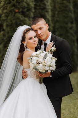 Siyah takım elbiseli şık bir damat ve uzun peçeli beyaz elbiseli şirin bir gelin eski bir evin yakınındaki bir parkta sarılıyorlar. Gülümseyen ve mutlu yeni evlilerin düğün portresi.