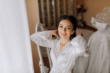 Saten bornozlu ve profesyonel makyajlı mutlu esmer gelin aynanın yanında duruyor ve düğün gününe hazırlanıyor.