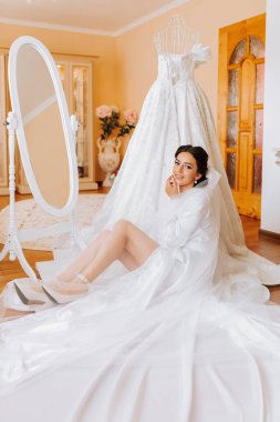 Uzun peçeli, profesyonel makyajlı, beyaz saten gecelikli mutlu esmer gelin. Gelin, aynanın yanındaki odada, yerde oturuyor. Bir mankenin üzerinde gelinlik.