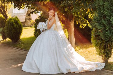 Sıcak güneşli bir gelin portresi. Elinde bir buket orkide, açık havada ve gün batımında. Sıcak yaz zamanı. Uzun bir peçe, konforlu beyaz bir elbise ve tren..