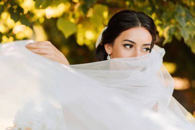 Sıcak güneşli bir gelin portresi. Elinde bir buket orkide, açık havada ve gün batımında. Sıcak yaz zamanı. Uzun bir peçe, konforlu beyaz bir elbise ve tren..