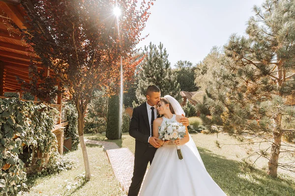 Siyah takım elbiseli şık bir damat ve uzun peçeli beyaz elbiseli şirin bir gelin eski bir evin yakınındaki bir parkta sarılıyorlar. Gülümseyen ve mutlu yeni evlilerin düğün portresi.