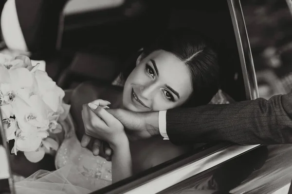 close-up portrait of a pretty shy bride in a car window, holding her groom\'s hand and looking at the camera