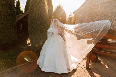 Sıcak güneşli bir gelin portresi. Elinde bir buket orkide, açık havada ve gün batımında. Sıcak yaz zamanı. Uzun bir peçe, konforlu beyaz bir elbise ve tren..