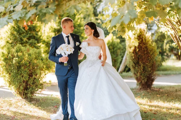結婚式の後にハッピーな花嫁とグルーミング ロングベール 開いた肩を持つシックな白いドレス オーケストラの花束 日没の間 — ストック写真