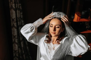 Sabahları uzun beyaz bir bornozla uzun siyah kıvırcık saçlı güzel bir gelin. Gelin düğün için hazırlanıyor. Profesyonel makyaj ve saç modeli. Başında bir taç ve gelinin uzun bir duvağı.