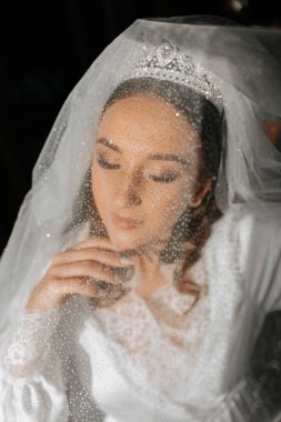 Sabahları uzun beyaz bir bornozla uzun siyah kıvırcık saçlı güzel bir gelin. Gelin düğün için hazırlanıyor. Profesyonel makyaj ve saç modeli. Başında bir taç ve gelinin uzun bir duvağı.