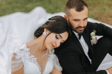 Modaya uygun bir damat ve beyaz elbiseli tatlı esmer gelin, damadın omzuna yaslanmış, parkta gülüyor çimenlerde, bahçede, dışarıda. Düğün fotoğrafı, gülümseyen yeni evlilerin portresi..