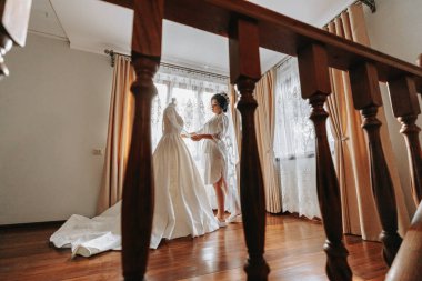 Mutlu bir gelin, güzel elbisesini elinde tutarak bir otel odasında lüks düğününe hazırlanıyor. Profesyonel saçlı, makyajlı ve sabahlık giymiş bir kadının portresi.