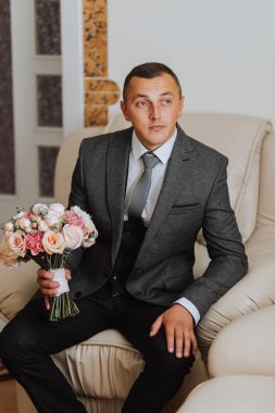 Portre. Beyaz gömlekli, klasik takım elbiseli bir adam koltukta oturuyor, elinde buketle poz veriyor. Şık bir saat. Erkek tarzı. Moda. İş dünyası