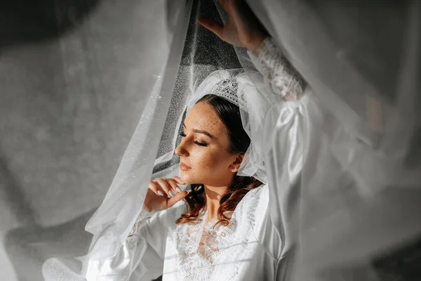 Beautiful bride with long dark curly hair in a long white robe in the morning. The bride is preparing for the wedding. Professional make-up and hair. A crown on the head and a long veil of the bride