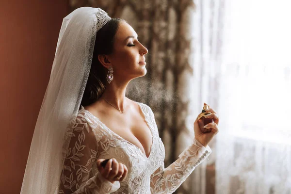 Female beauty. Cute woman at home. The bride sprays perfume on her body. A stylish woman wears a white dress. Spray delicate perfume. Stylish glass bottle of perfume in hands.